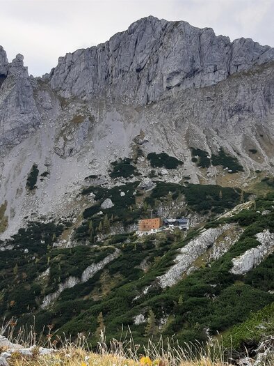 Eine beeindruckende Berglandschaft mit steilen Felsen und grünen Hügeln. Im Hintergrund ist ein kleines Gebäude zu sehen.
