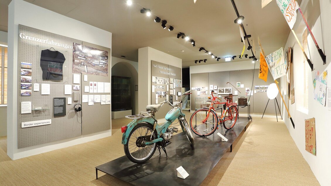 Ein Ausstellungsraum mit zwei Fahrrädern, einem blauen und einem roten, sowie verschiedenen Informationsmaterialien und Bildern an den Wänden. Der Raum ist gut beleuchtet und zeigt historische Elemente. | © Universalmuseum Joanneum - J. Kucek +