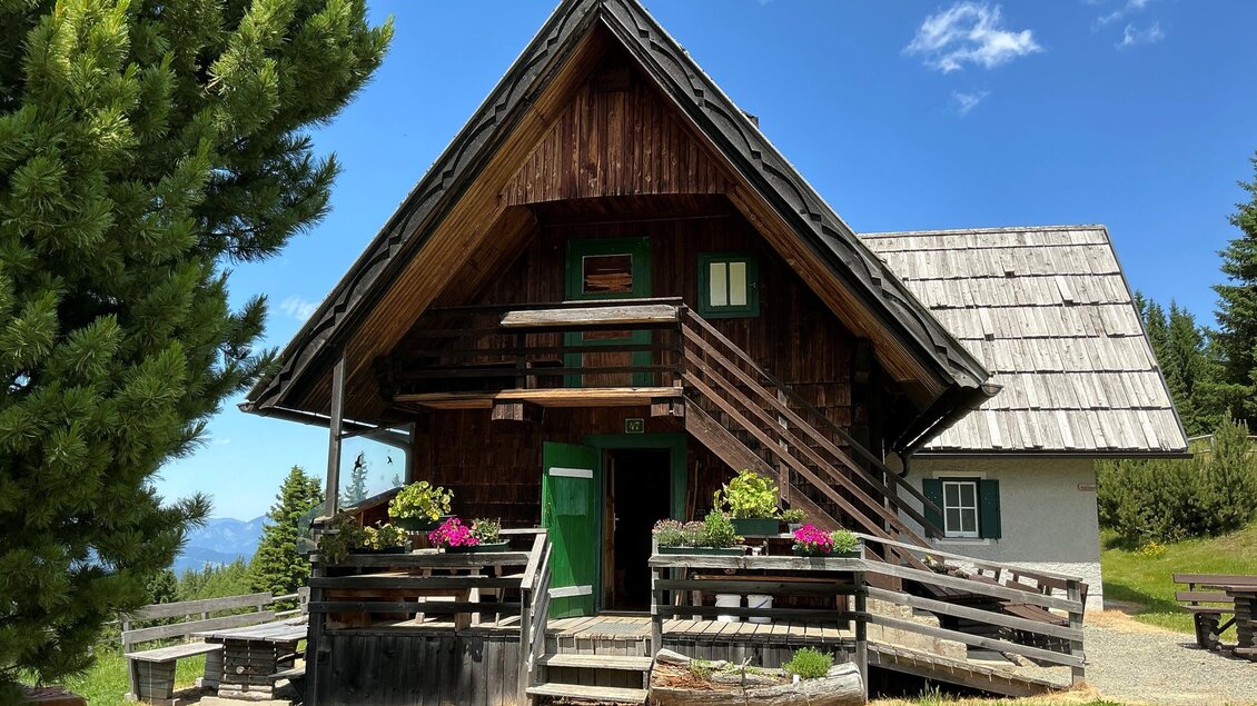 Ein charmantes Holzhaus mit einem schrägen Dach, umgeben von grünen Wiesen und Bäumen. Blumen blühen auf der Veranda und der Himmel ist blau mit einigen Wolken. | © Margareta Köstner