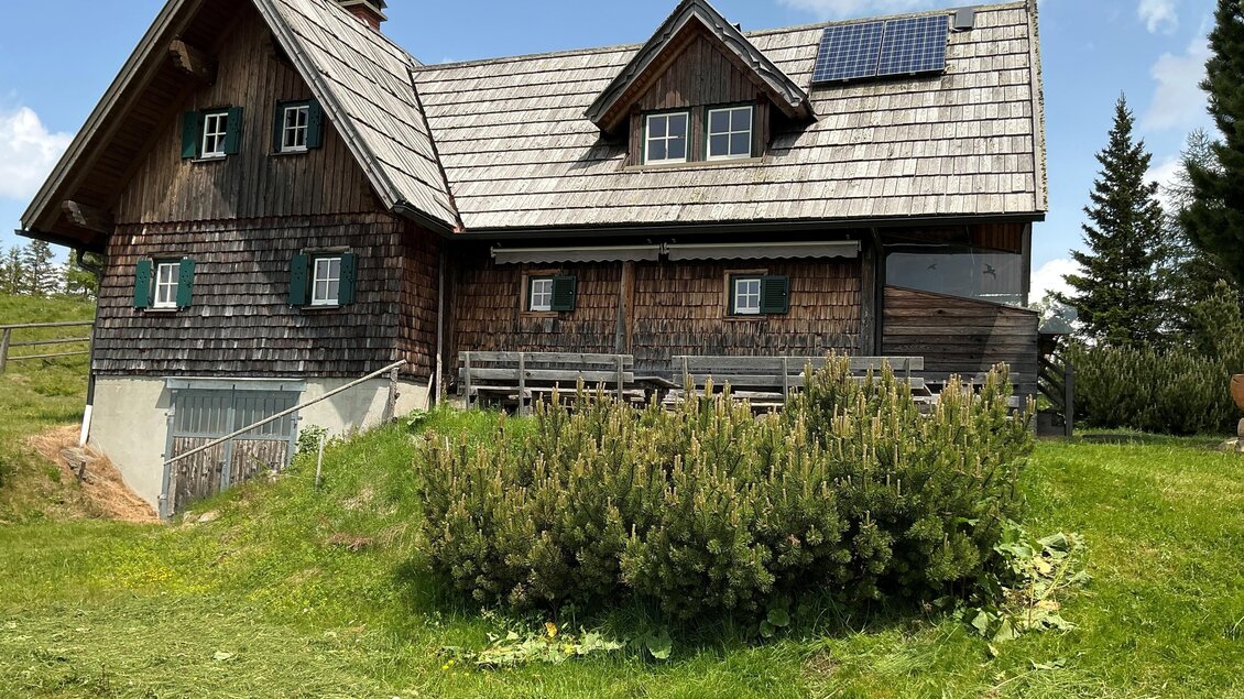 Ein rustikales Holzhaus mit einem grazilen Dach und Solarpanels. Umgeben von grünem Gras und Büschen, strahlt es ländlichen Charme aus. | © Margareta Köstner