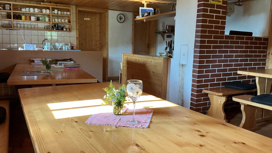 Ein gemütlicher Innenraum mit Holztischen und einem dekorativen Tisch mit einer kleinen Blumenvase. Im Hintergrund sind Regale mit Geschirr und ein Kamin zu sehen. | © Margareta Köstner