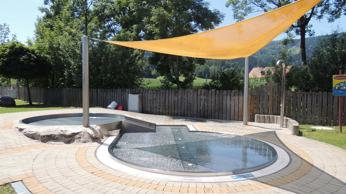 Ein entspannender Außenbereich mit einem runden Wasserbecken und einer gelben Sonnenschutzplane. Im Hintergrund sind Bäume und eine umzäunte Fläche zu sehen. | © WIP Erlebnisbad Wernersdorf