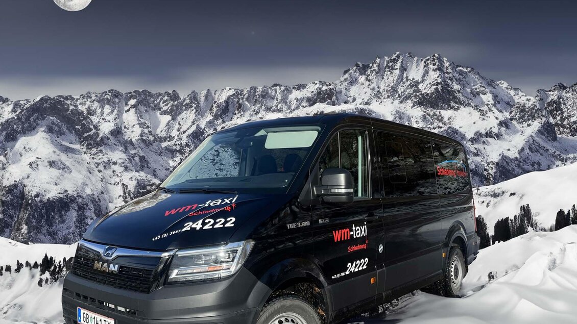 Ein schwarzer Kleinbus steht in einer schneebedeckten Landschaft vor majestätischen Bergen. Der Himmel ist klar und der Mond leuchtet hell. | © WM Taxi Schladming