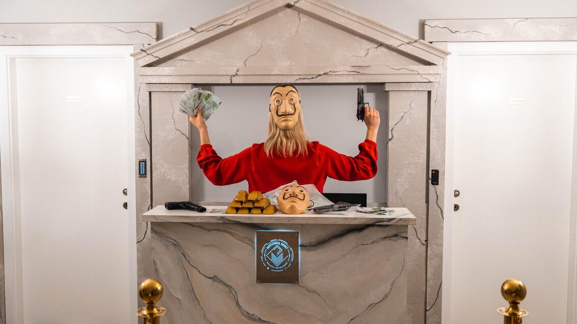 Person mit Maske und Requisiten wie Geld, Goldbarren und Spielzeugpistole hinter Bankschalter | © WOOP!