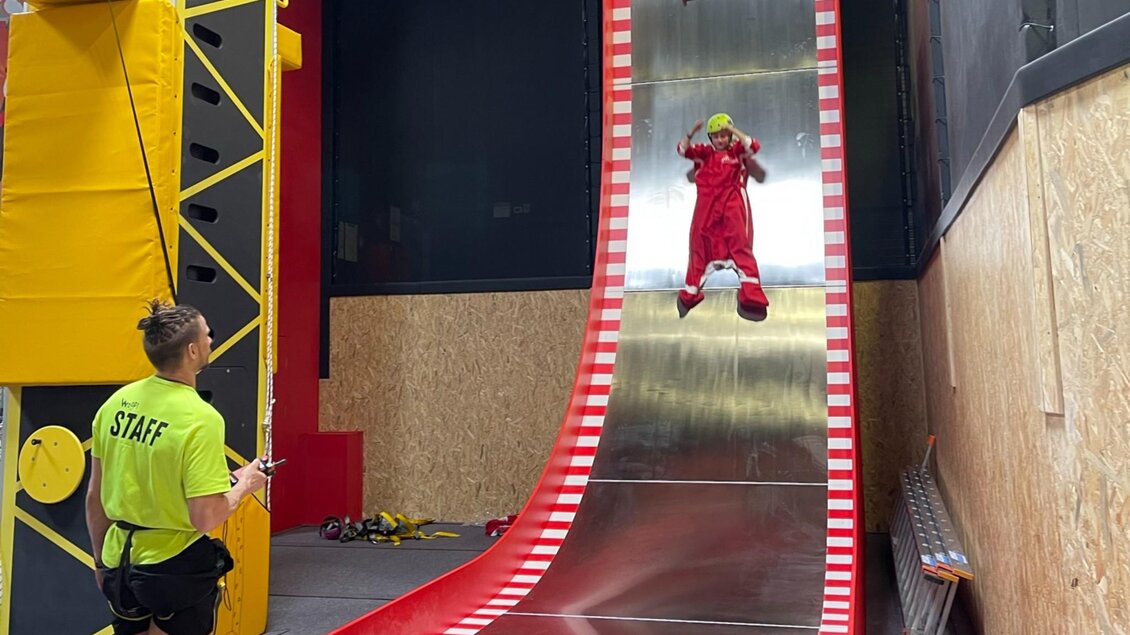 Kind im Overall mit Helm rutscht senkrecht eine hohe Metallrampe hinunter, betreut von Mitarbeiter | © WOOP!