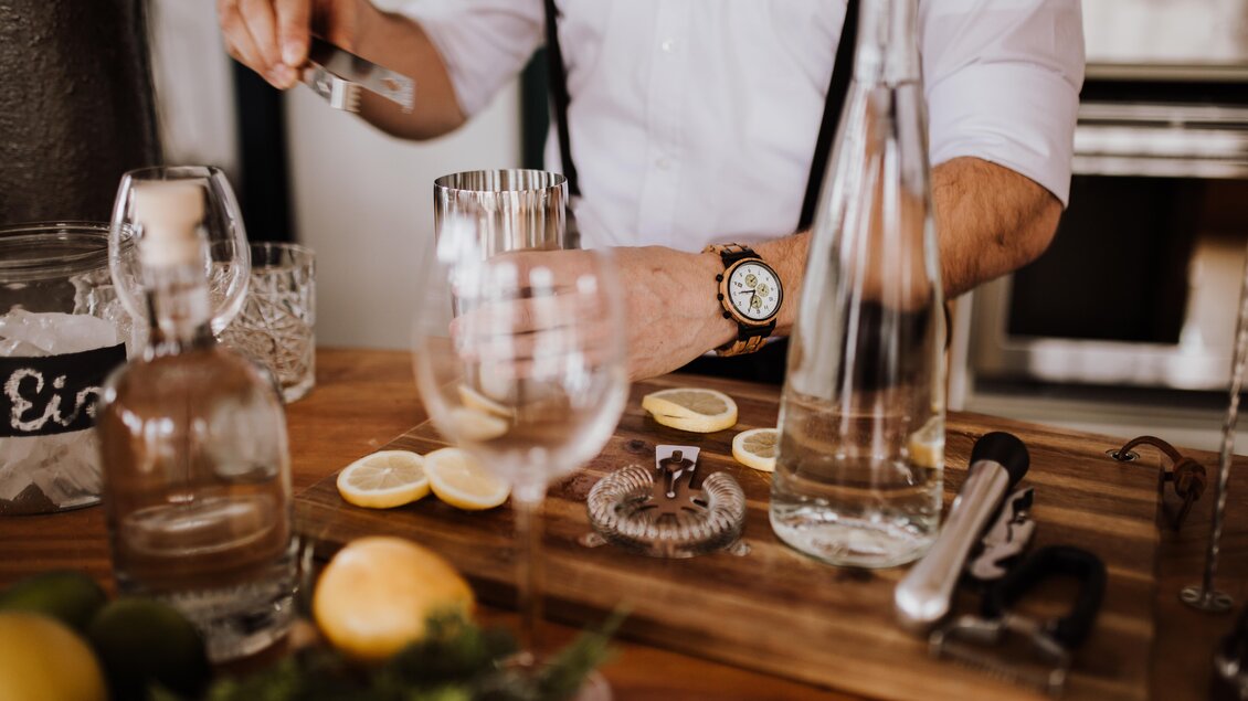 Ein Bartender mixt Cocktails mit frischen Zitronen und Limetten auf einem Holztisch. Im Hintergrund sind Gläser und Spirituosen zu sehen. | © Mira Schaffer