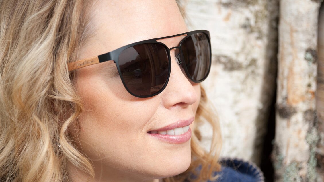 Eine Frau trägt stylische Sonnenbrillen und lächelt. Im Hintergrund sind Baumrinden sichtbar. | © Waidzeit