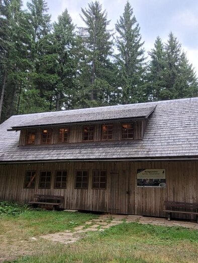 A wooden house in a forest area, surrounded by tall trees. The sky is cloudy and the terrain is green and inviting. | © TV Südsteiermark - Irene Löschnig