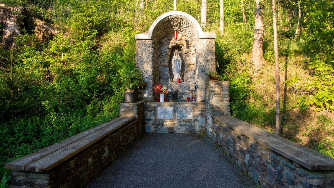 Eine kleine Kapelle aus Stein mit einer Statue stehen inmitten eines Waldgebiets. Vor der Kapelle befinden sich Bänke und Blumen in einem ruhigen, natürlichen Umfeld. | © Kurkommission Bad Blumau