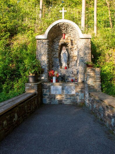 A small stone chapel with a statue stands in the middle of a forest area. In front of the chapel are benches and flowers in a tranquil, natural setting. | © Kurkommission Bad Blumau