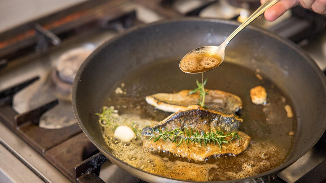 Ein Koch brät frischen Fisch in einer Pfanne mit aromatischen Kräutern. Eine Hand gibt eine Flüssigkeit mit einem Löffel hinzu. | © Netzwerk Kulinarik wildbild