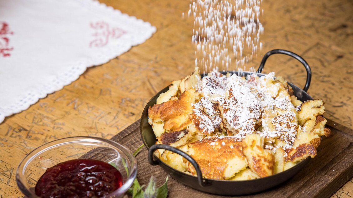 Ein köstlicher Kaiserschmarrn wird mit Puderzucker bestreut und serviert. Dazu gibt es eine Schale mit roter Marmelade. | © Netzwerk Kulinarik wildbild