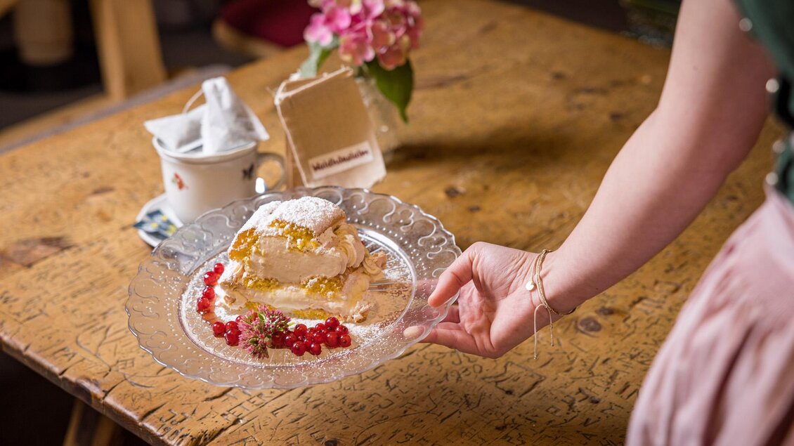 Ein Stück Torte auf einem schönen Teller, serviert von einer Hand. Im Hintergrund steht eine Blume in einer Vase auf einem Holztisch. | © Netzwerk Kulinarik wildbild