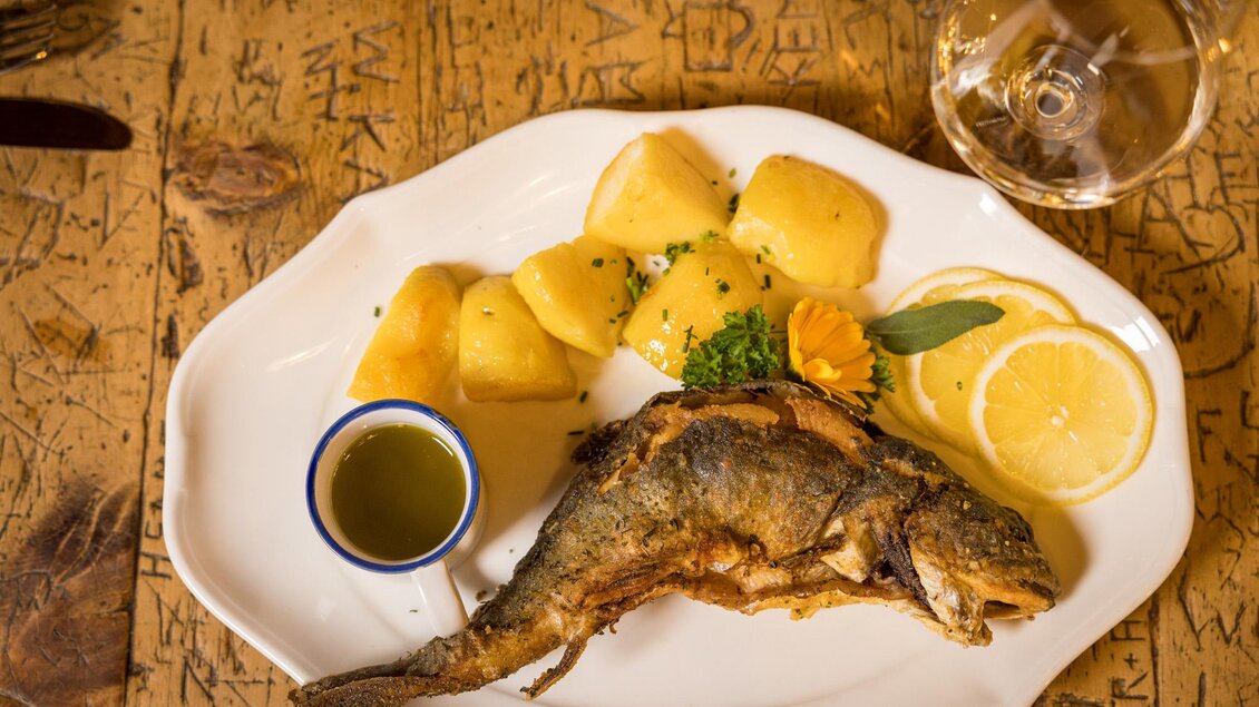 Ein gebratener Fisch auf einem weißen Teller, serviert mit Kartoffeln, Zitronenscheiben und einer kleinen Schale mit Olivenöl. Im Hintergrund steht ein Glas Wasser. | © Netzwerk Kulinarik wildbild