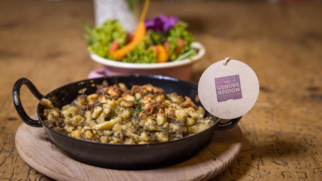 Ein köstliches Gericht in einer kleinen Pfanne, überbacken mit knusprigen Brötchenstücken. Im Hintergrund ein frischer Salat mit buntem Gemüse. | © Netzwerk Kulinarik wildbild