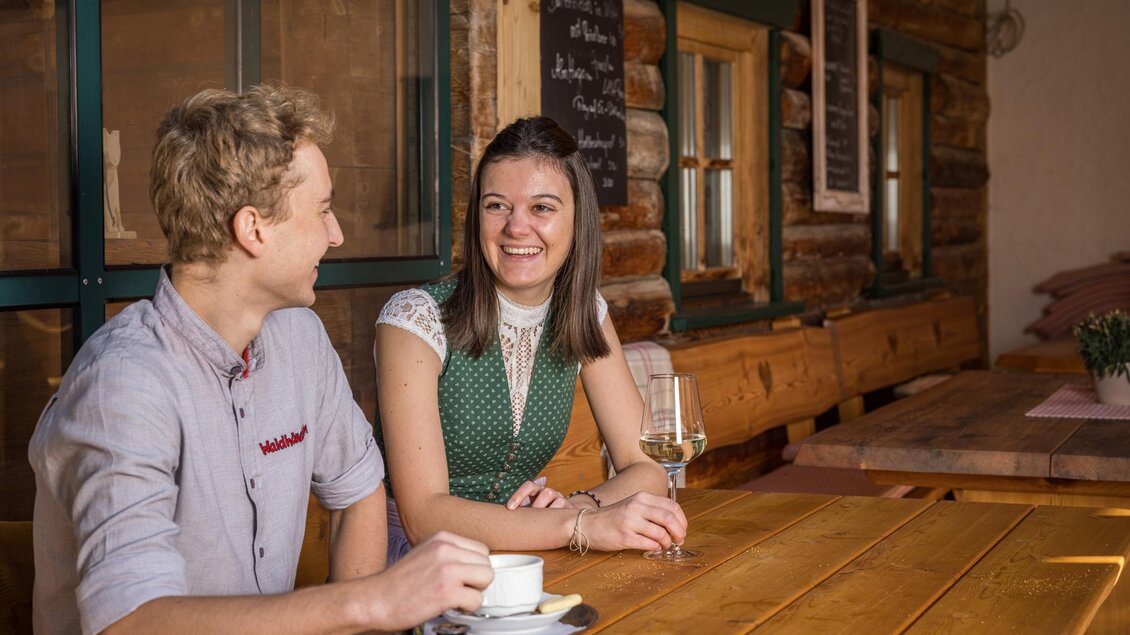 Zwei Menschen sitzen an einem Holztisch in einem gemütlichen Restaurant. Sie lächeln und genießen gemeinsame Zeit bei einem Getränk. | © Netzwerk Kulinarik wildbild
