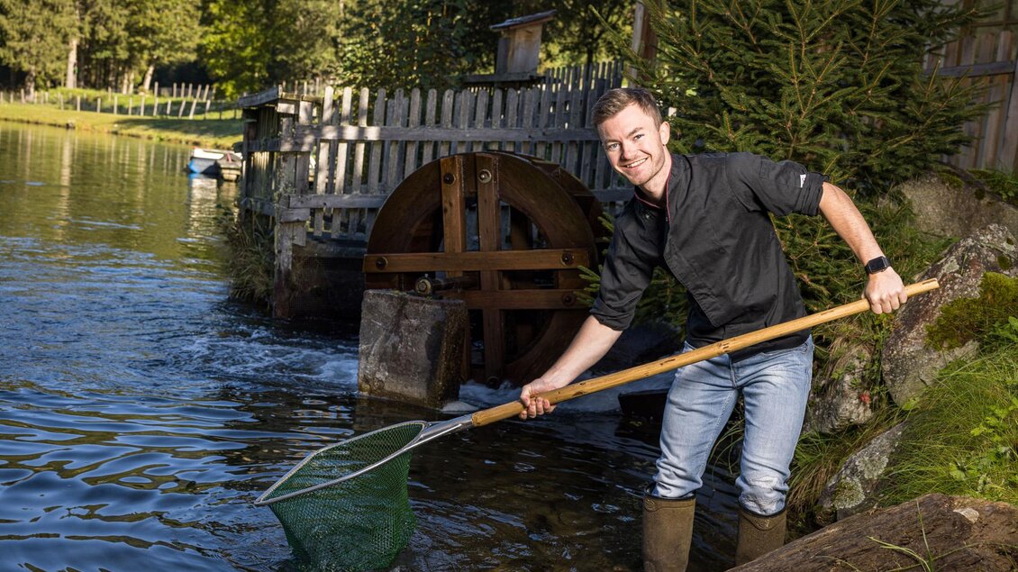 Ein Mann steht am Wasser und hält einen Kescher in der Hand. Im Hintergrund ist ein Wasserrad und viel grüne Vegetation zu sehen. | © Netzwerk Kulinarik wildbild
