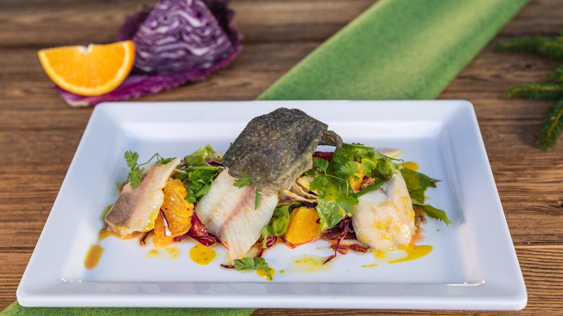 Ein kunstvolles Gericht mit frischem Fisch, buntem Salat und Orangenscheiben. Das Gericht wird auf einem weißen Teller serviert und hat ansprechende Farben. | © Martin Huber