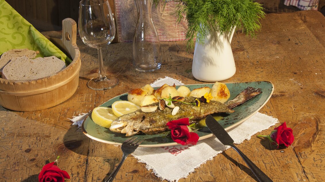 Ein köstlicher gebratener Fisch auf einem Teller, garniert mit Zitrone und Kräutern. Neben dem Fisch liegen Kartoffeln und frische Rosen auf dem Tisch. | © Waldhäuslalm