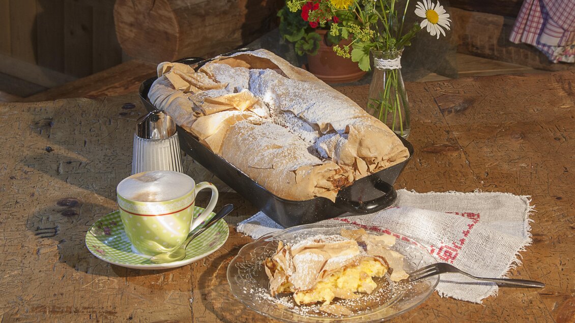 Ein frisch gebackener Kuchen auf einem rustikalen Tisch, neben einer Tasse Kaffee. Im Hintergrund stehen bunte Blumen in einer Vase. | © Waldhäuslalm