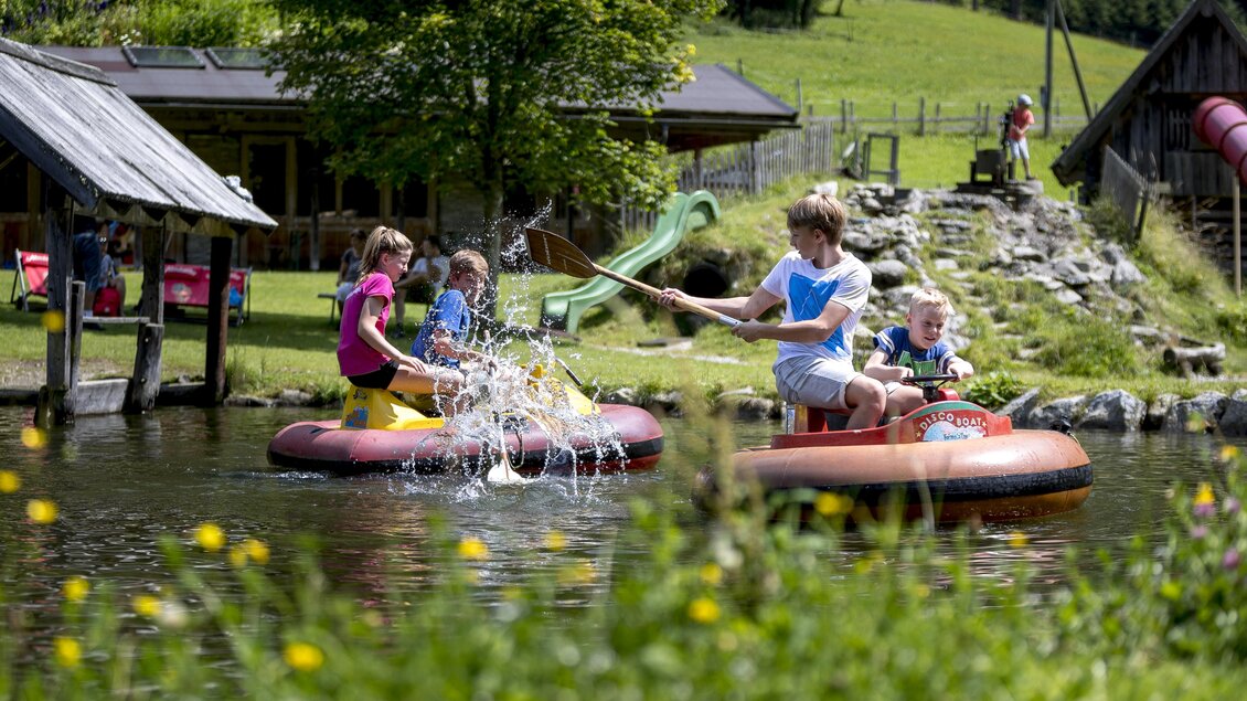 Kinder haben Spaß beim Bootfahren auf einem ruhigen Teich. Im Hintergrund sind Bäume und eine Spielanlage zu sehen. | © Tom Lamm