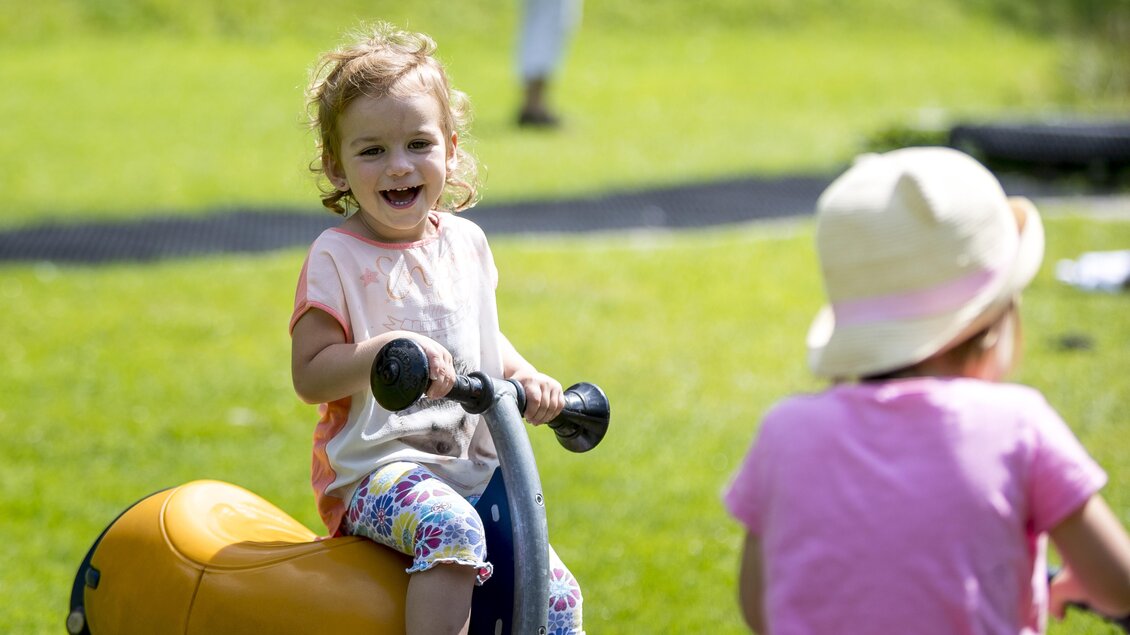Ein kleines Mädchen fährt fröhlich auf einem Spielzeug und lächelt. Im Hintergrund spielt ein weiteres Kind auf der Wiese. | © Tom Lamm