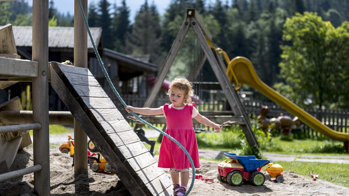 Ein kleines Mädchen in einem pinken Kleid spielt auf einem Spielplatz. Im Hintergrund gibt es eine Rutsche und verschiedene Spielzeuge. | © Tom Lamm