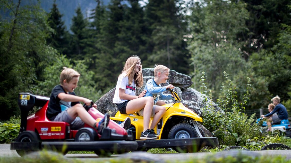 Kinder fahren fröhlich auf kleinen Elektroautos in der Natur. Im Hintergrund sind Bäume und eine entspannte Atmosphäre zu sehen. | © Tom Lamm