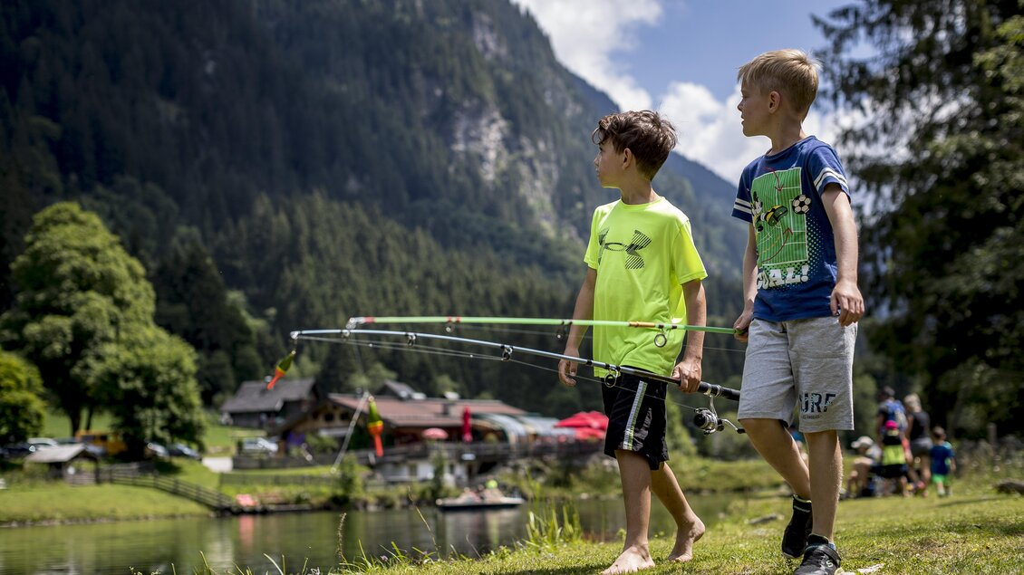 Zwei Kinder gehen am Ufer eines ruhigen Sees spazieren und halten Angelruten in den Händen. Im Hintergrund sind grüne Wälder und Häuser zu sehen. | © Tom Lamm