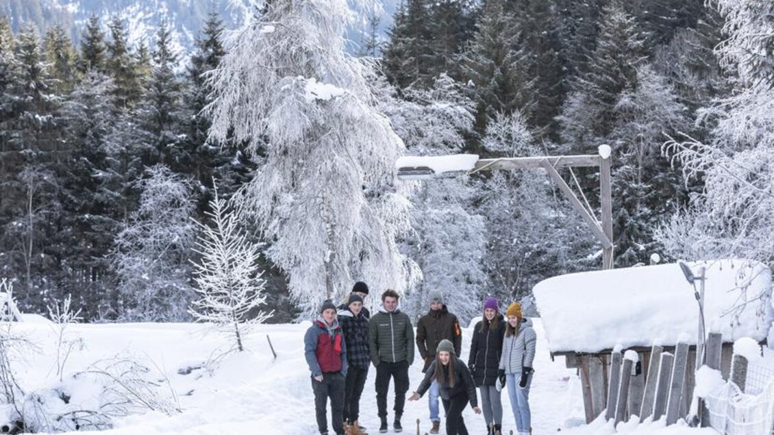 Eine Gruppe von sechs Personen spielt auf einem zugefrorenen Gewässer in einer winterlichen Landschaft. Im Hintergrund sind schneebedeckte Berge und frostige Bäume zu sehen. | © Waldhäuslalm/Martin Huber