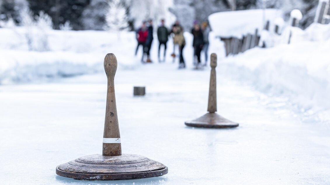Zwei Holzplatten zum Eisstockschießen stehen auf einer vereisten Fläche. Im Hintergrund sind Personen zu sehen, die die winterliche Szenerie genießen. | © Waldhäuslalm/Martin Huber