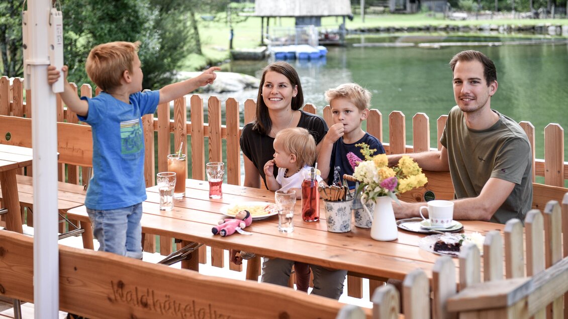 Eine Familie sitzt an einem Holztisch am Wasser. Kinder spielen und die Atmosphäre ist entspannt und freundlich. | © Waldhäuslalm/Shootingstar