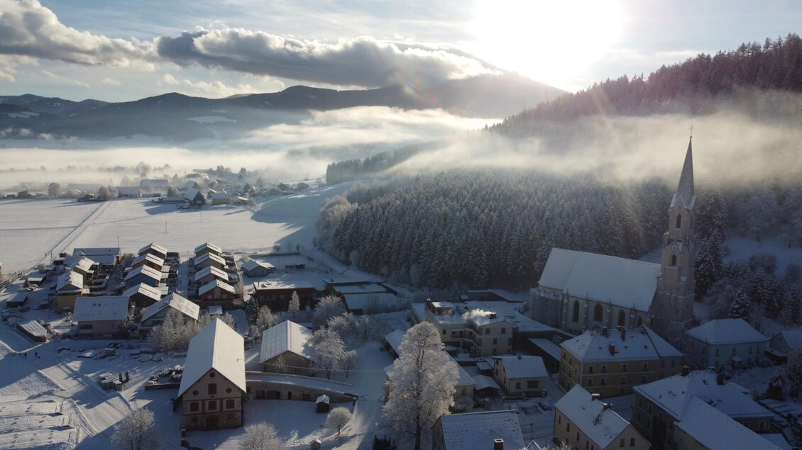 Eine malerische Winterlandschaft mit verschneiten Häusern und sanften Hügeln. Die Sonne scheint über dem Dorf und erzeugt eine warme, goldene Atmosphäre. | © Diazöse Graz Seckau