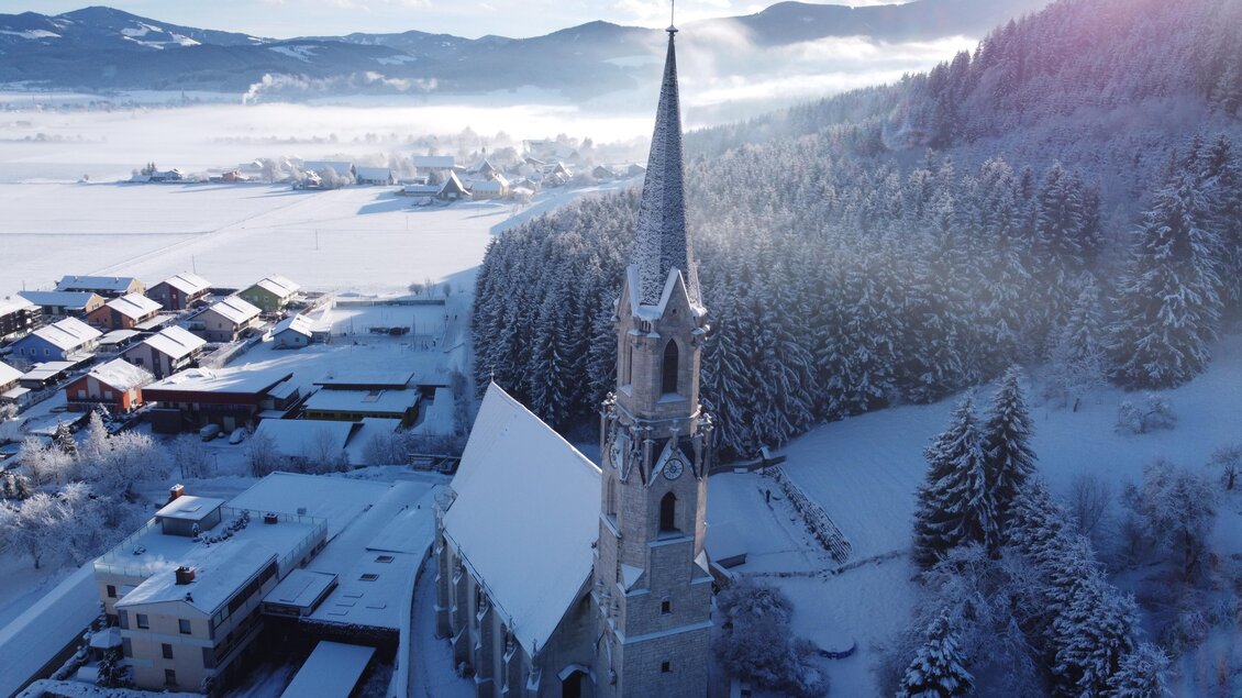 Eine schneebedeckte Landschaft mit einer malerischen Kirche und einem hohen Kirchturm. Im Hintergrund sind bewaldete Hügel und ein klarer Himmel zu sehen. | © Diazöse Graz Seckau