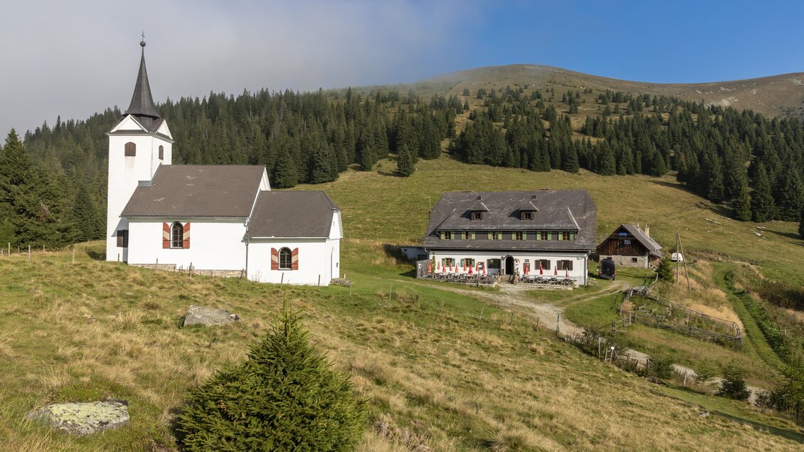 Eine malerische Landschaft mit einer weißen Kirche und einem Bauernhaus. Im Hintergrund sind sanfte Hügel und ein blauer Himmel zu sehen. | © TV Region Graz - Lunghammer