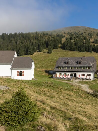 Eine malerische Landschaft mit einer weißen Kirche und einem Bauernhaus. Im Hintergrund sind sanfte Hügel und ein blauer Himmel zu sehen. | © TV Region Graz - Lunghammer