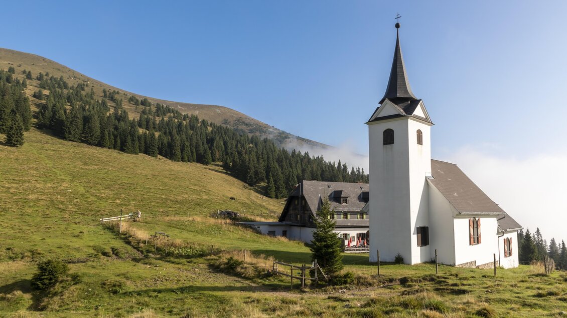 Eine weiße Kirche mit spitzem Turm steht in einer grünen Landschaft. Im Hintergrund sind bewaldete Hügel und ein klarer Himmel zu sehen. | © TV Region Graz Lunghammer