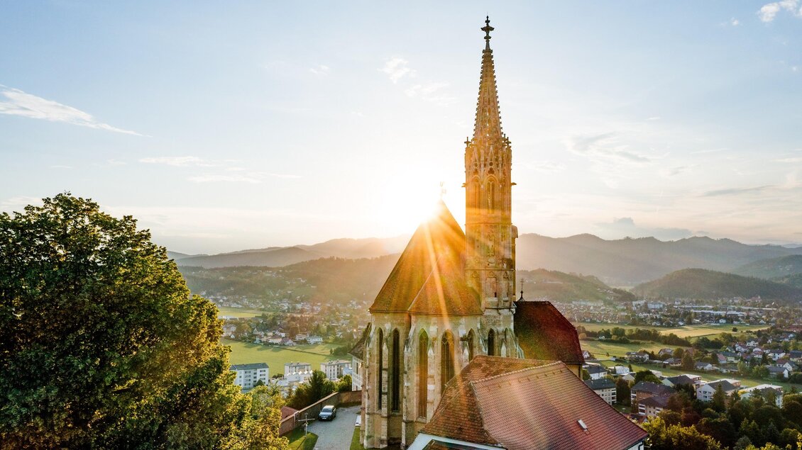 Eine Kirche mit einem hohen Turm steht im Vordergrund. Die Sonne geht am Horizont unter und beleuchtet die Landschaft im Hintergrund. | © Region Graz - Mias Photoart
