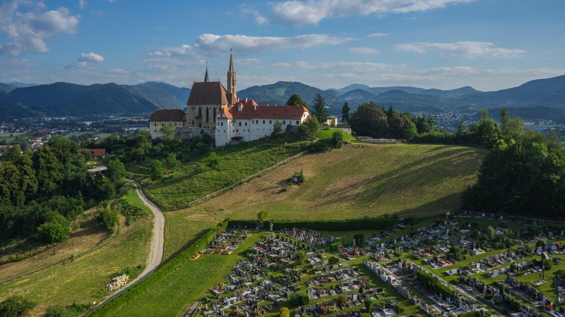 Eine beeindruckende Kirche steht auf einem Hügel, umgeben von einem gepflegten Friedhof und grünen Feldern. Im Hintergrund sind sanfte Berge und ein blauer Himmel zu sehen. | © TV Region Graz - picfly