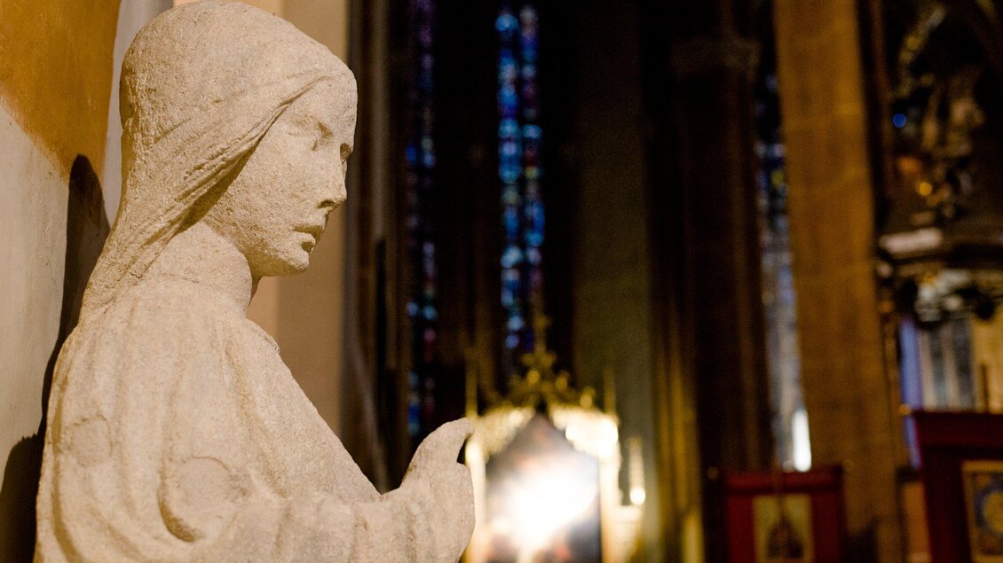 Eine Statue einer nachdenklich wirkenden Figur in einer Kirche. Im Hintergrund sind farbige Buntglasfenster zu sehen. | © TV Region OberGraz-Mias Photoart