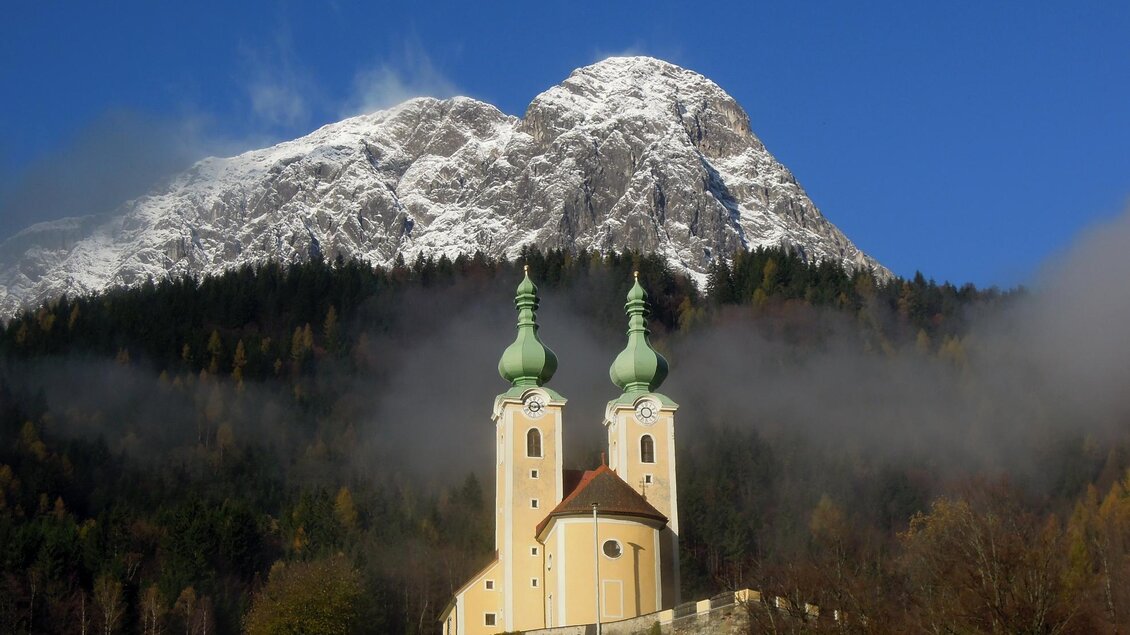 Eine malerische Kirche mit zwei Türmen steht vor einem schneebedeckten Berg. Der Himmel ist klar und blau, umgeben von leichtem Nebel und grünen Wäldern. | © TV ERZBERG LEOBEN
