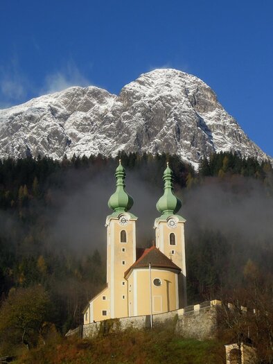 A picturesque church with two towers stands in front of a snow-covered mountain. The sky is clear and blue, surrounded by light fog and green forests. | © TV ERZBERG LEOBEN