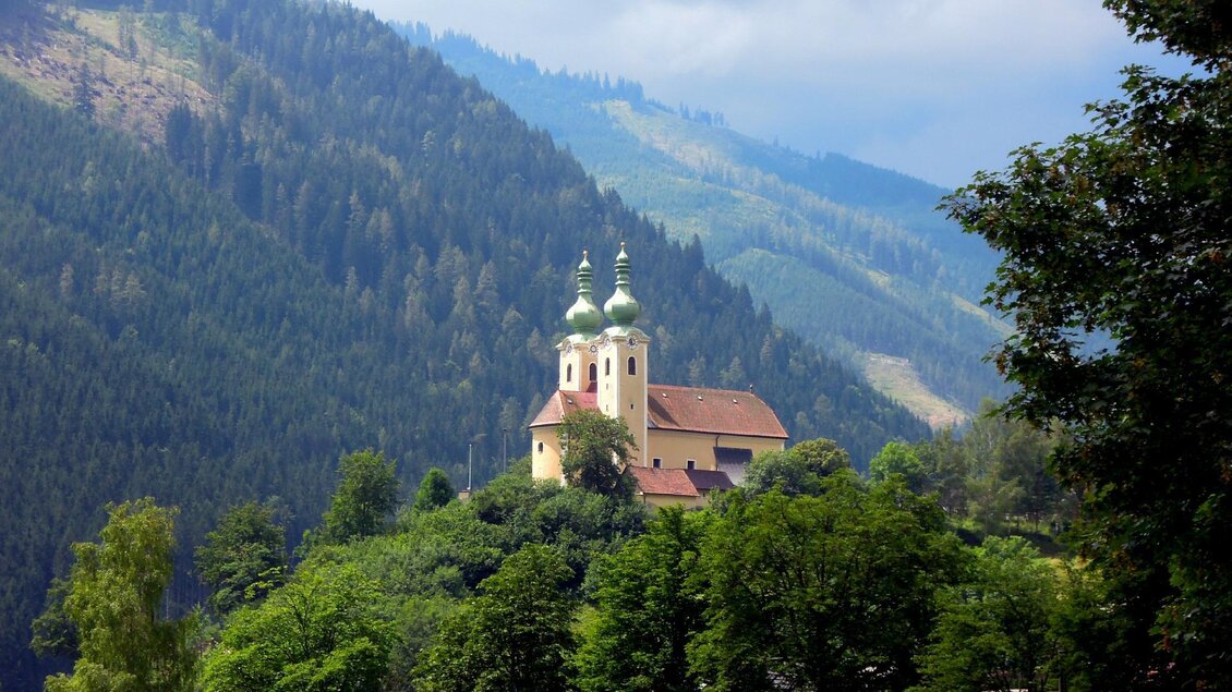 Eine malerische Kirche steht auf einem Hügel, umgeben von grünen Bäumen. Im Hintergrund sind schneebedeckte Berge und ein bewölkter Himmel zu sehen. | © TV ERZBERG LEOBEN