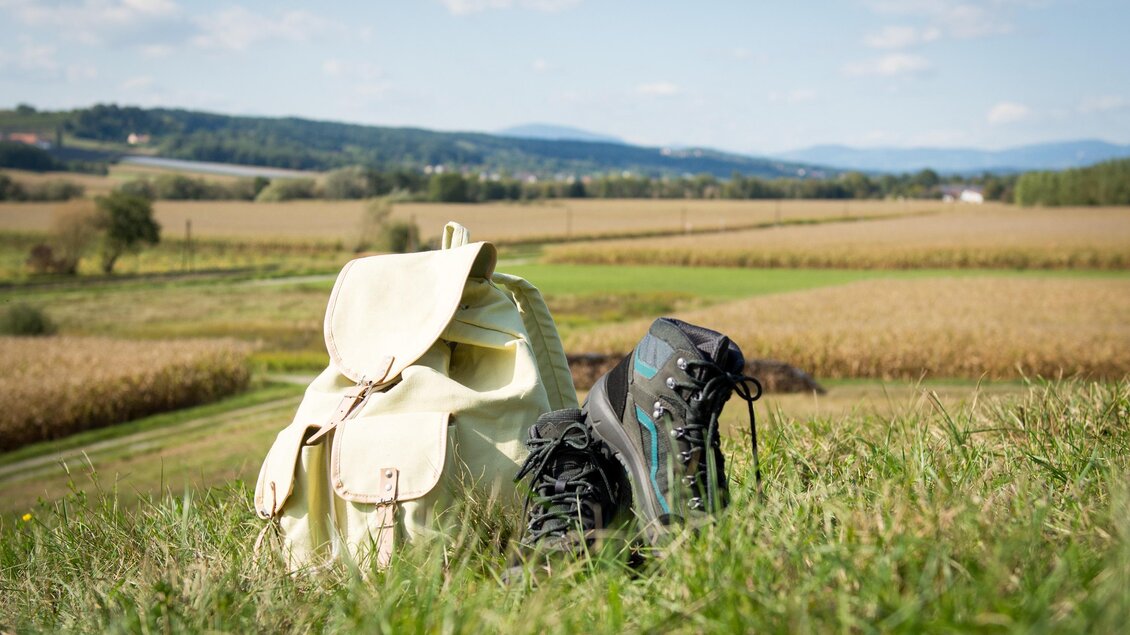 Ein Rucksack und ein Paar Wanderschuhe stehen auf einer grünen Wiese. Im Hintergrund erstreckt sich eine weite Landschaft mit Feldern und Bergen. | © Kurkommission Bad Blumau