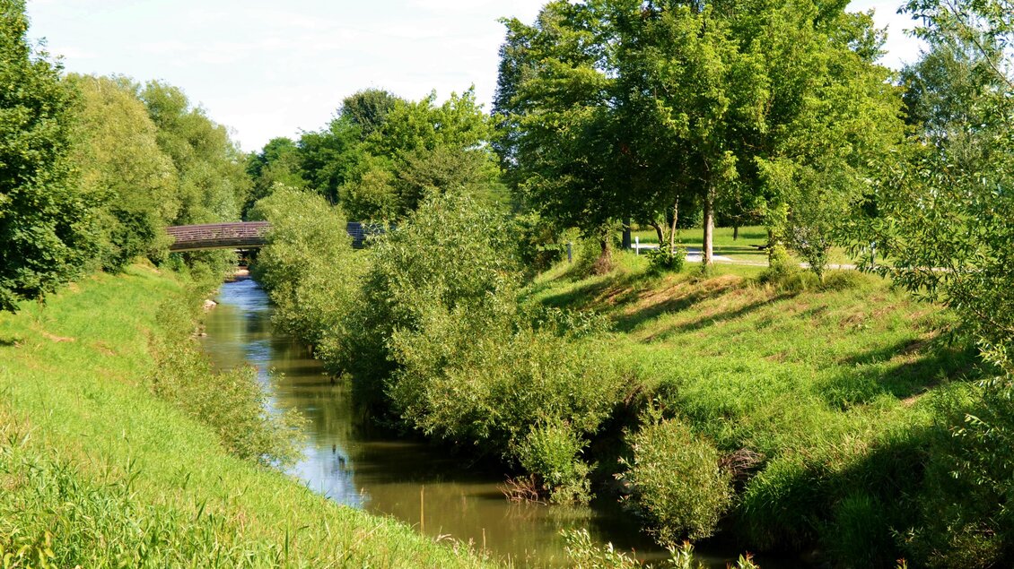 Ein ruhiger Fluss fließt durch eine grüne Landschaft mit Bäumen und Sträuchern. Im Hintergrund ist eine Holzbrücke zu sehen. | © Kurkommission Bad Blumau