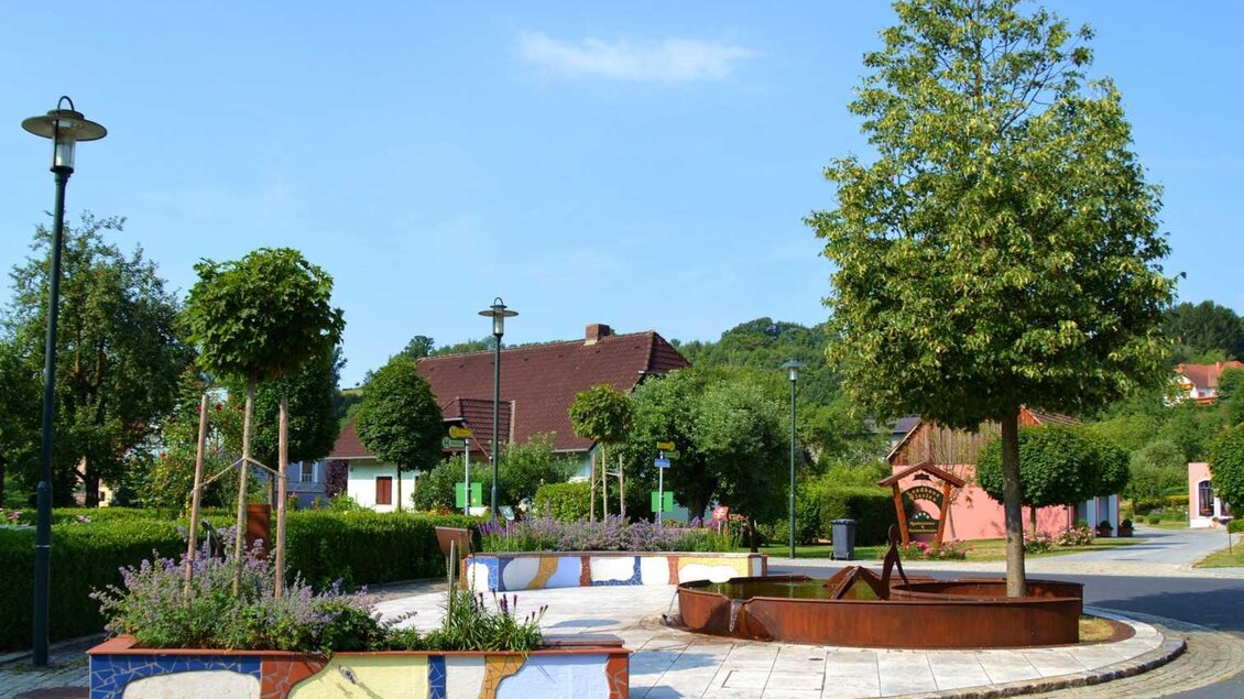 Eine bunte Grünanlage mit Blumenbeeten und Bäumen in einer ruhigen Straße. Im Hintergrund sind einige Häuser und Laternen sichtbar. | © Kurkommission Bad Blumau