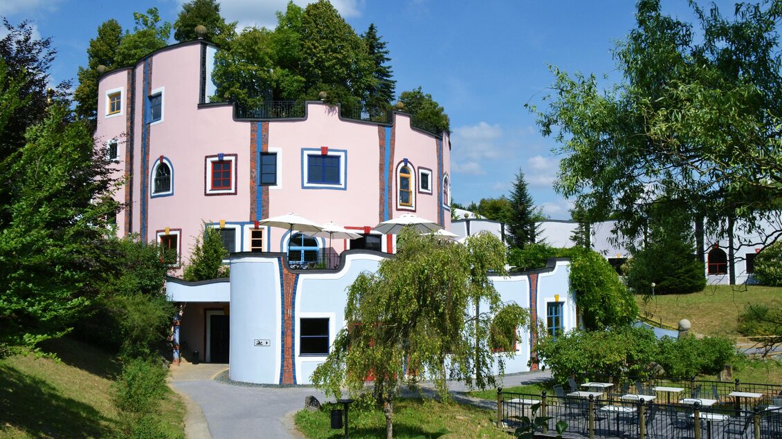 Ein modernes, farbenfrohes Gebäude mit runden Formen und großen Fenstern, umgeben von grünen Bäumen. Der Garten ist gepflegt und bietet Plätze zum Entspannen. | © Kurkommission Bad Blumau