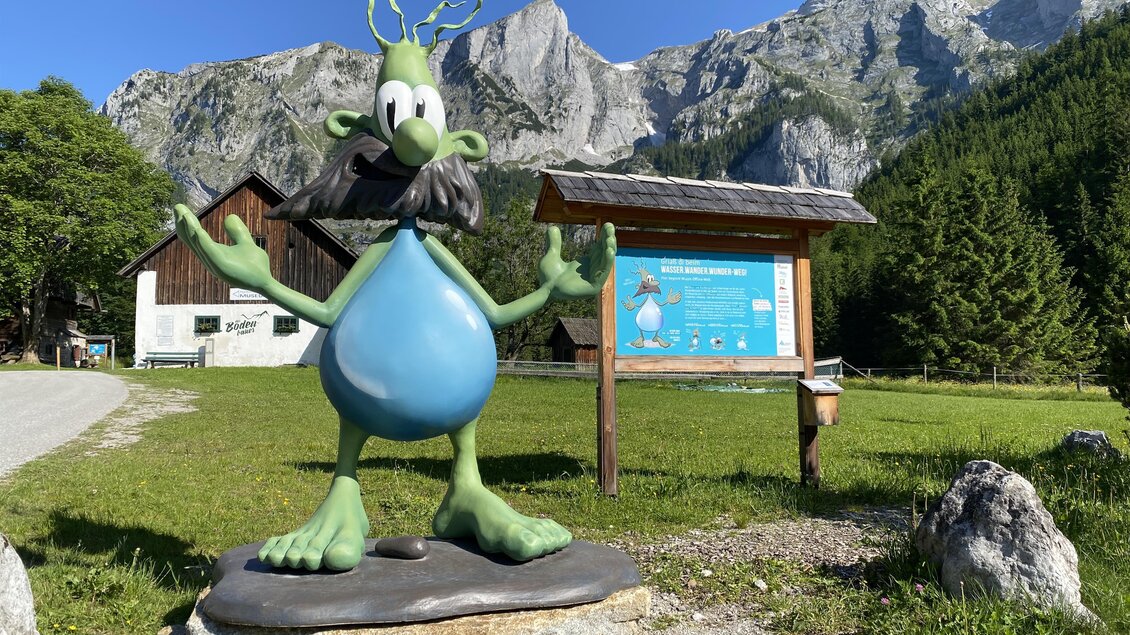 Eine bunte Statue eines Cartoon-Charakters steht in einer malerischen Berglandschaft. Im Hintergrund sind hohe Berge und ein Informationsschild sichtbar.