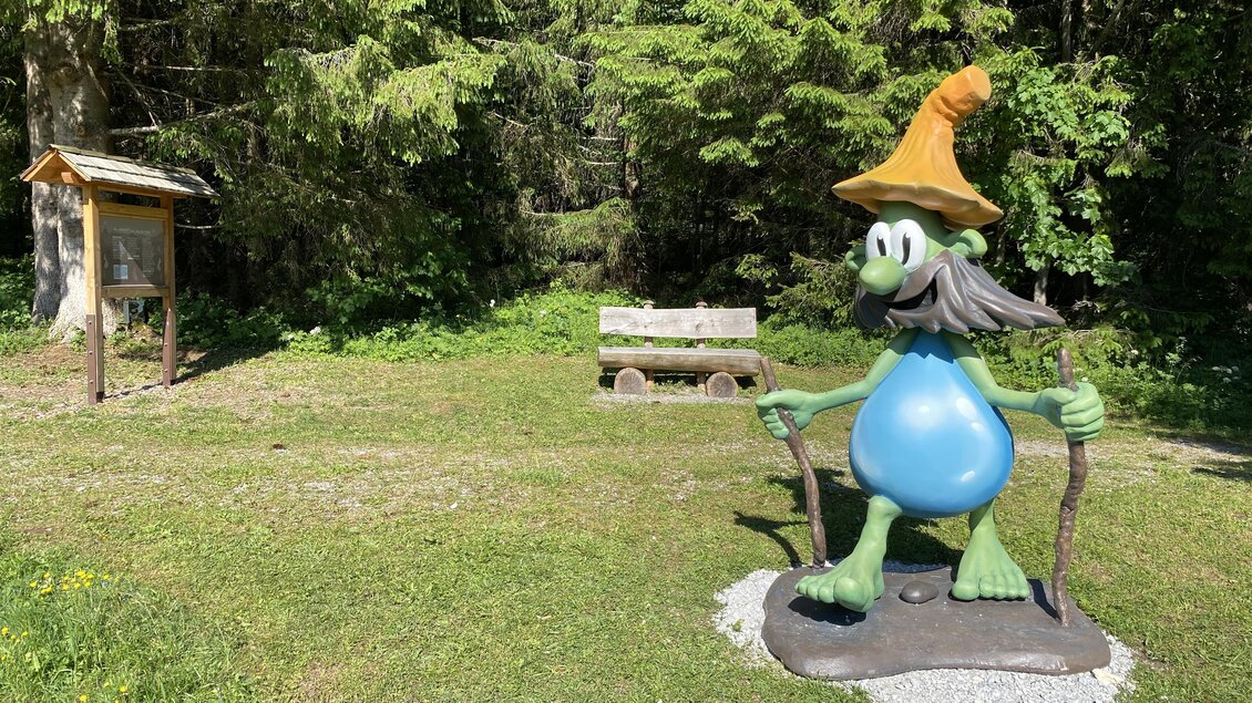 Eine bunte Skulptur eines Trolls steht in einer grünen Umgebung. Im Hintergrund sind Holzbank und ein Informationsstand zu sehen.