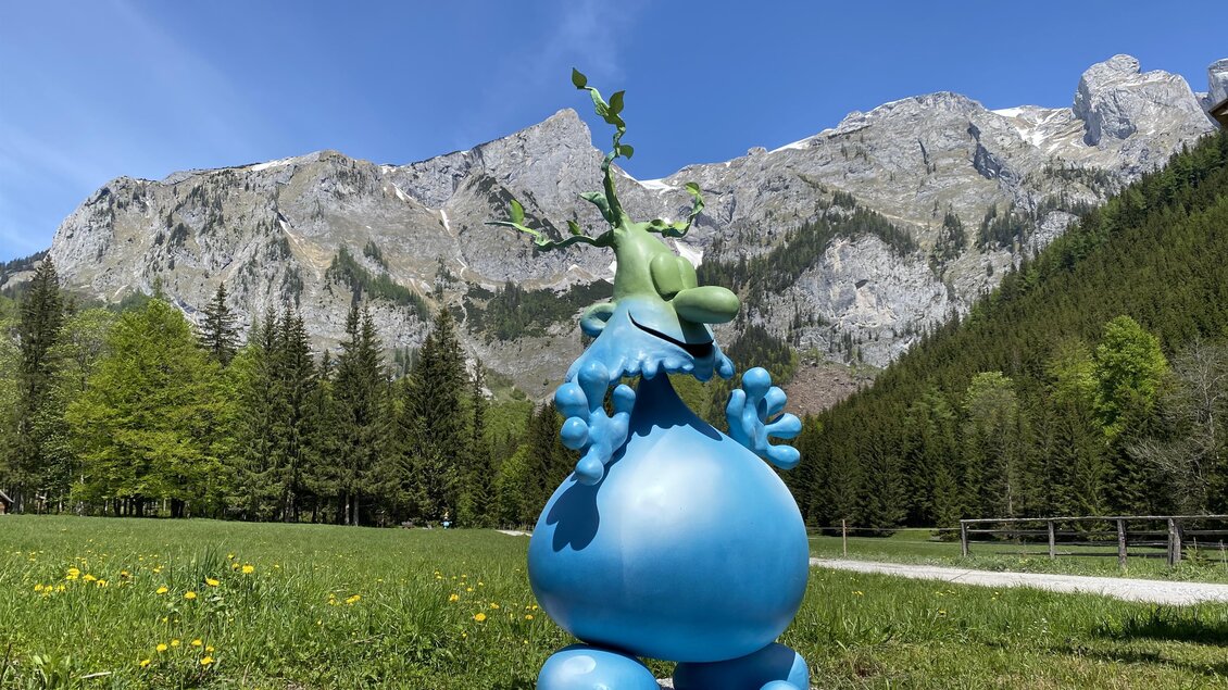 Eine bunte Statue eines Comic-Charakters steht auf einer Wiese mit Bergen im Hintergrund. Die Statue hat grüne Blätter auf dem Kopf und ist von einer schönen Landschaft umgeben.